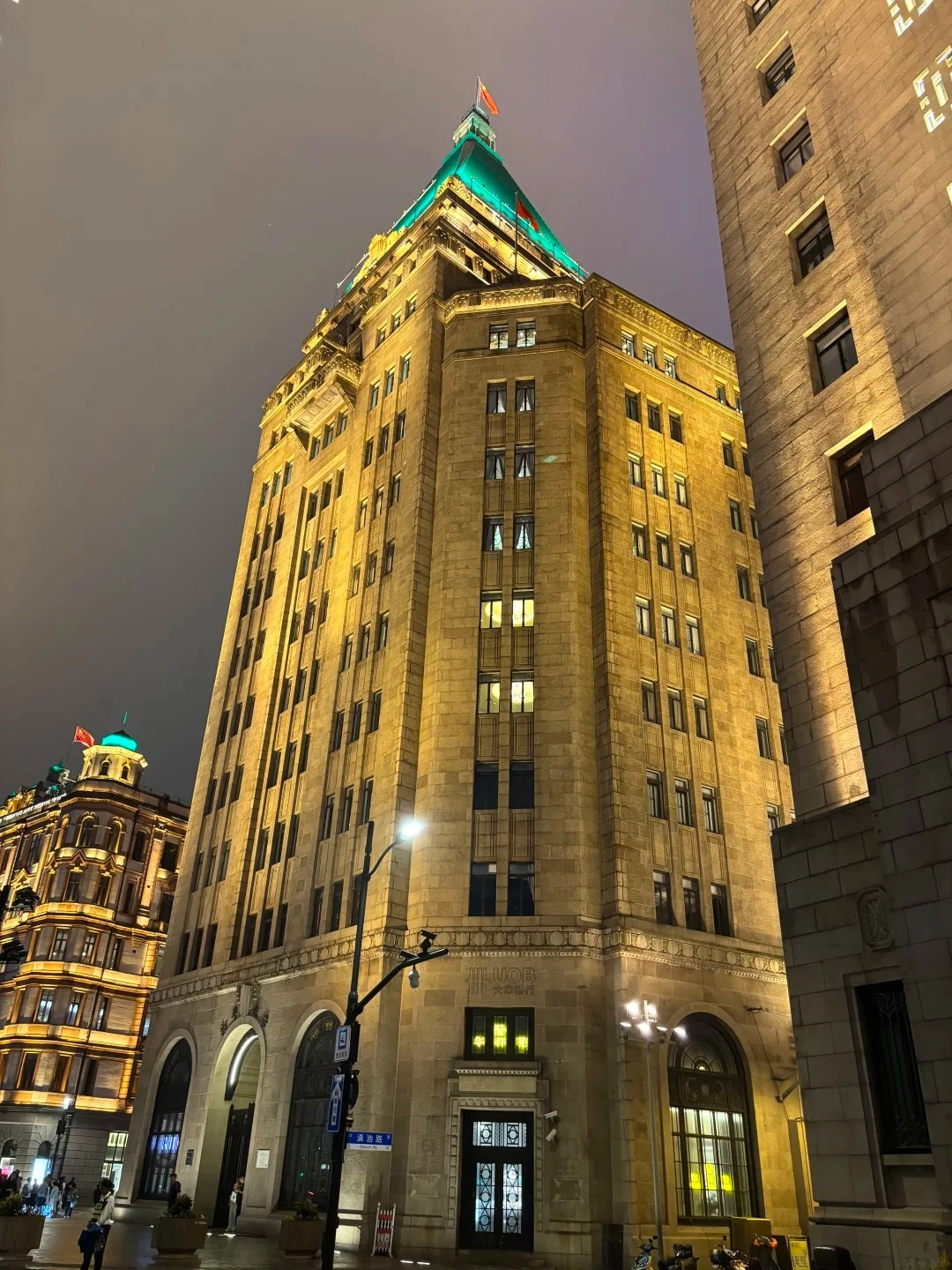 A tall, golden-lit Art Deco building with a distinctive green pyramidal roof topped with a small red flag, viewed from a low angle at twilight. Another lighter-colored building stands to its right, and a similar building with ornate detailing and a green dome is visible to the left and further back. The street in front is dimly lit by streetlights and shows a few pedestrians and parked motorcycles.