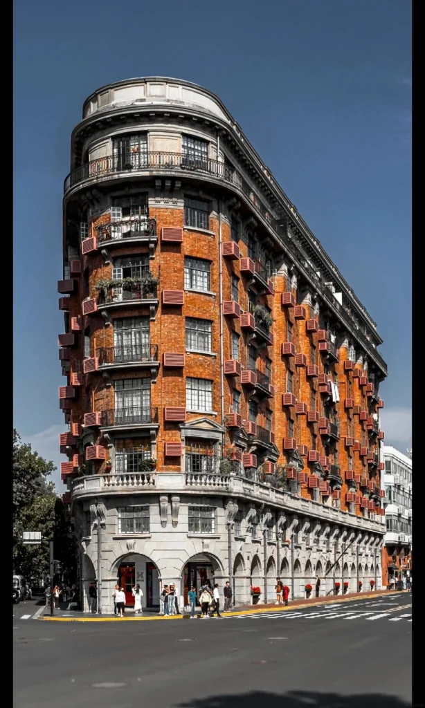 A wide shot of the Wukang Mansion, a prominent Art Deco building in Shanghai, under a clear blue sky. The building has a distinctive curved facade, constructed of red brick with numerous windows and balconies. The lower floors feature arched entrances and gray stone detailing. Several people are visible walking on the street in front of the building.