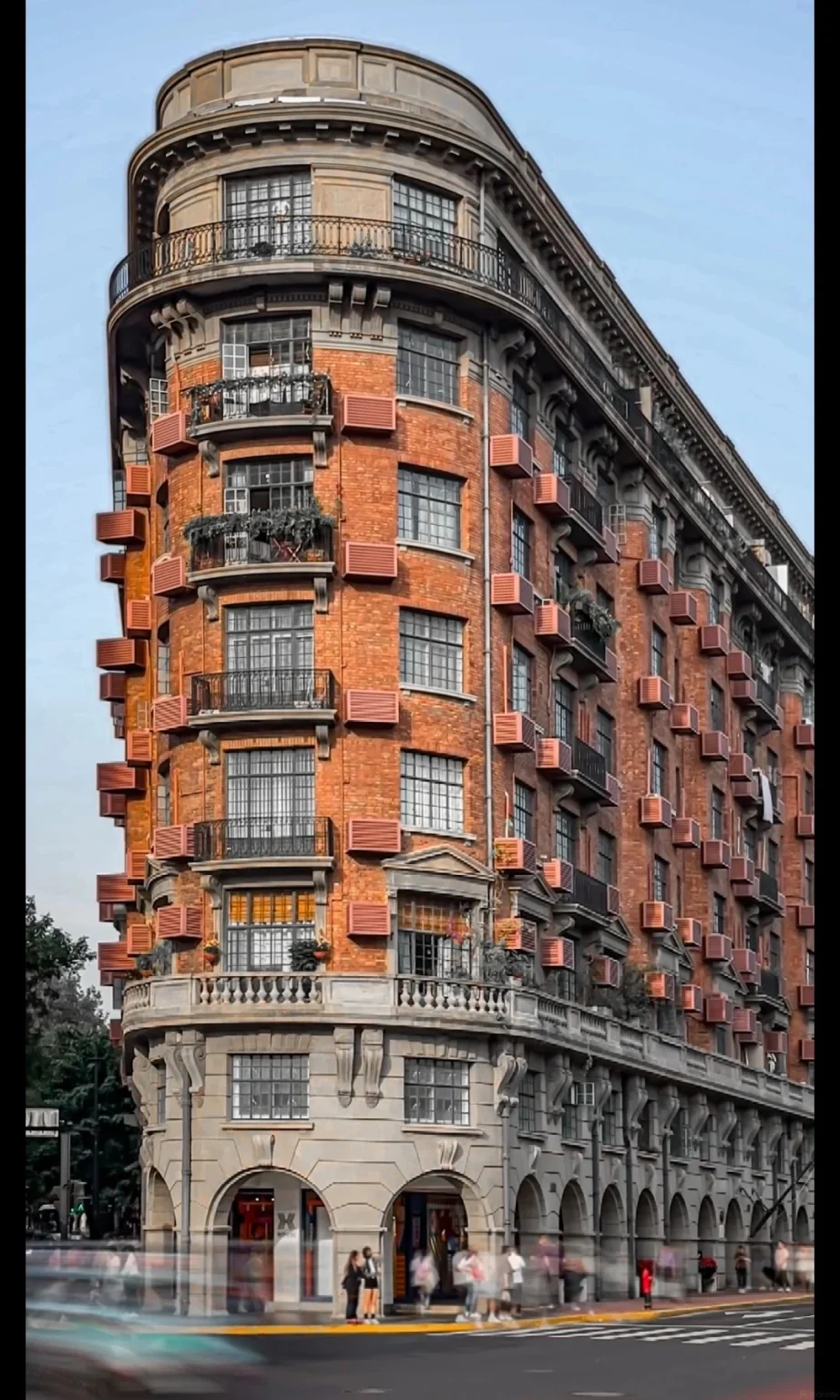 A curved, multi-story Art Deco building made of red brick with numerous balconies, grey stone arcades at the base, and small, red-slatted window shades on many floors.