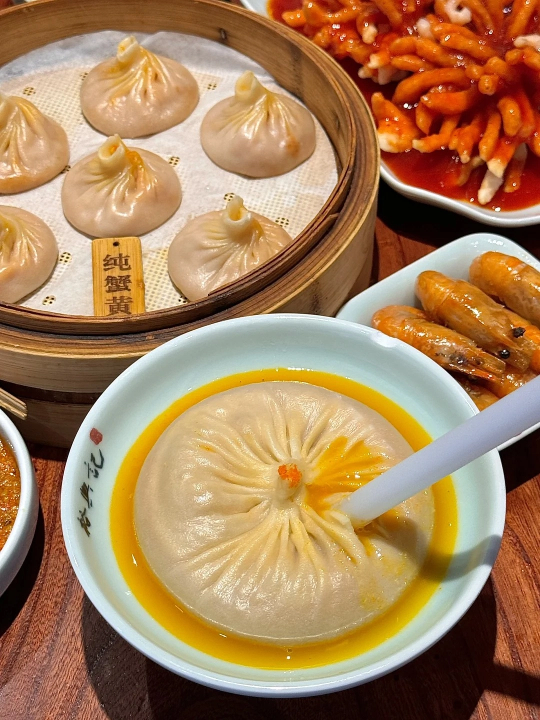 A large soup dumpling, known as xiaolongbao, sits in a shallow bowl of broth, with a white straw inserted into it. Several more steamed soup dumplings are arranged in a bamboo steamer basket in the background, along with a plate of shrimp and a dish of what appears to be red braised pork or chicken.