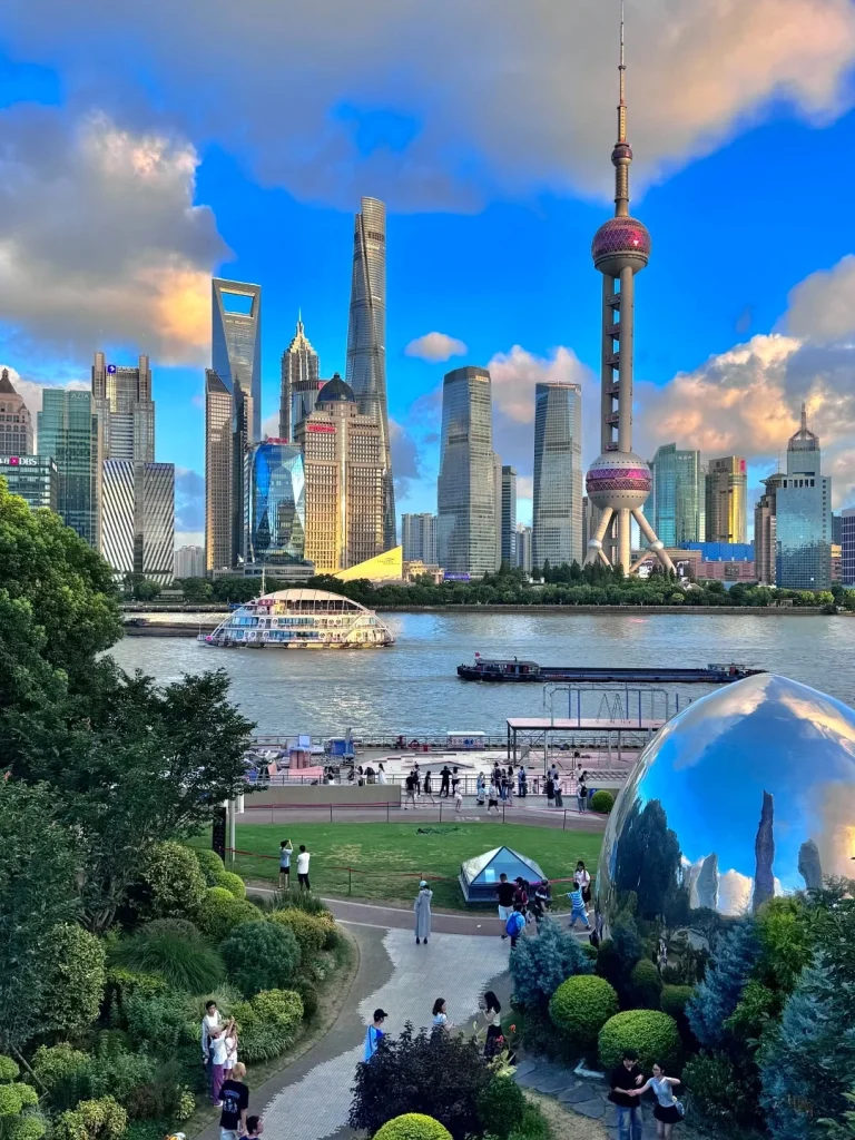 A wide shot of the Lujiazui skyline in Shanghai, China, viewed from a green park along the Huangpu River. In the foreground, lush bushes and a winding path lead to a large, reflective, egg-shaped sculpture. People are walking and gathered in the park. Across the river, the Pudong skyline features prominent skyscrapers including the Oriental Pearl Tower, Shanghai Tower, and Jin Mao Tower. A large, multi-decked boat is on the river, with smaller boats and barges also visible. The sky is a vibrant blue with scattered white clouds, and the sunlight casts a warm glow on the buildings and landscape.