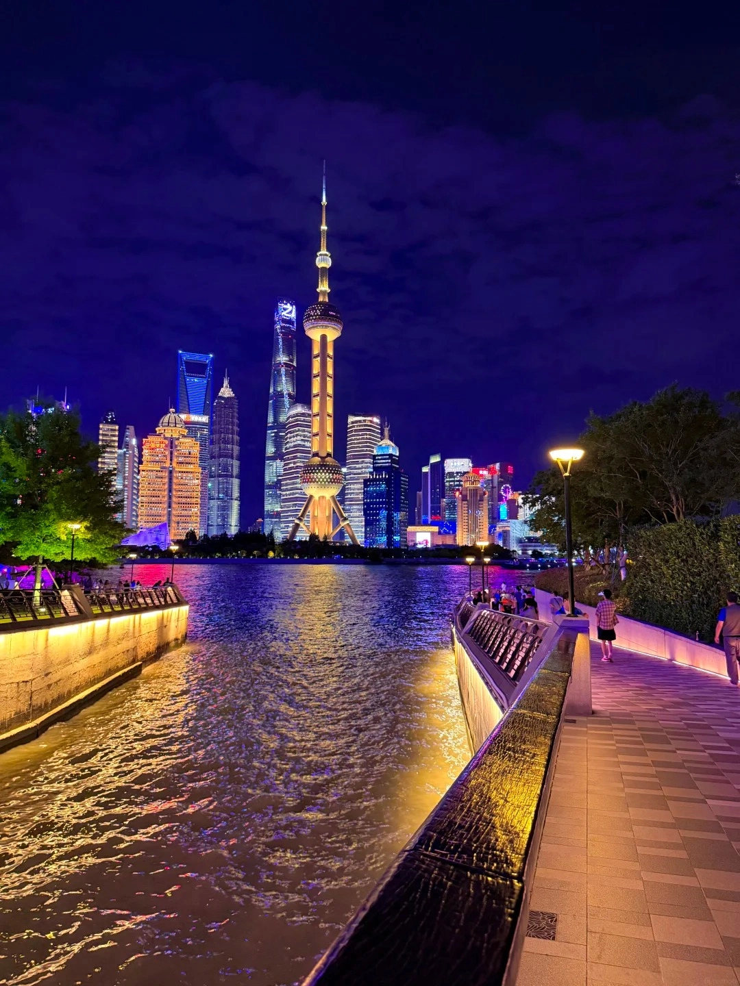 A nighttime view of the Shanghai skyline with the Oriental Pearl Tower and other skyscrapers illuminated, viewed from a riverfront walkway with people strolling and colorful lights reflecting on the water.