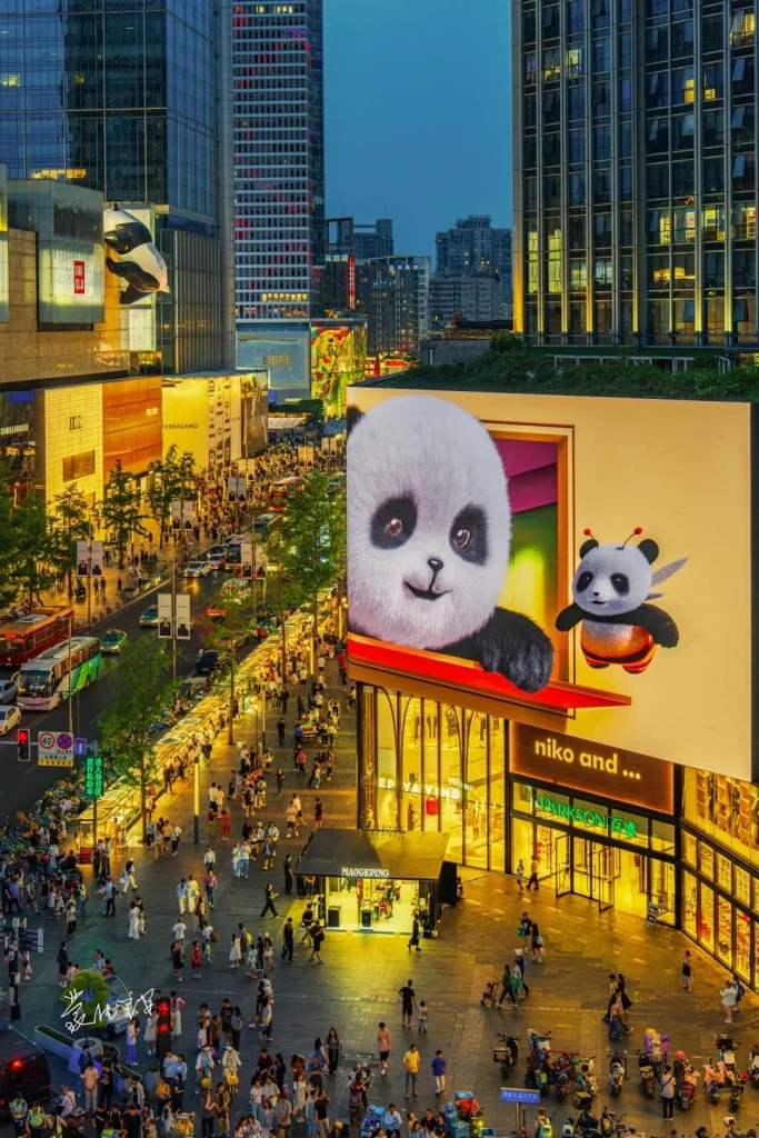 A wide shot of a busy pedestrian street at dusk, lined with modern buildings and illuminated storefronts. A large billboard features a giant cartoon panda and a smaller bee-panda. People are walking on the street, and cars and buses are visible on the road. The sky is a deep blue, suggesting evening.
