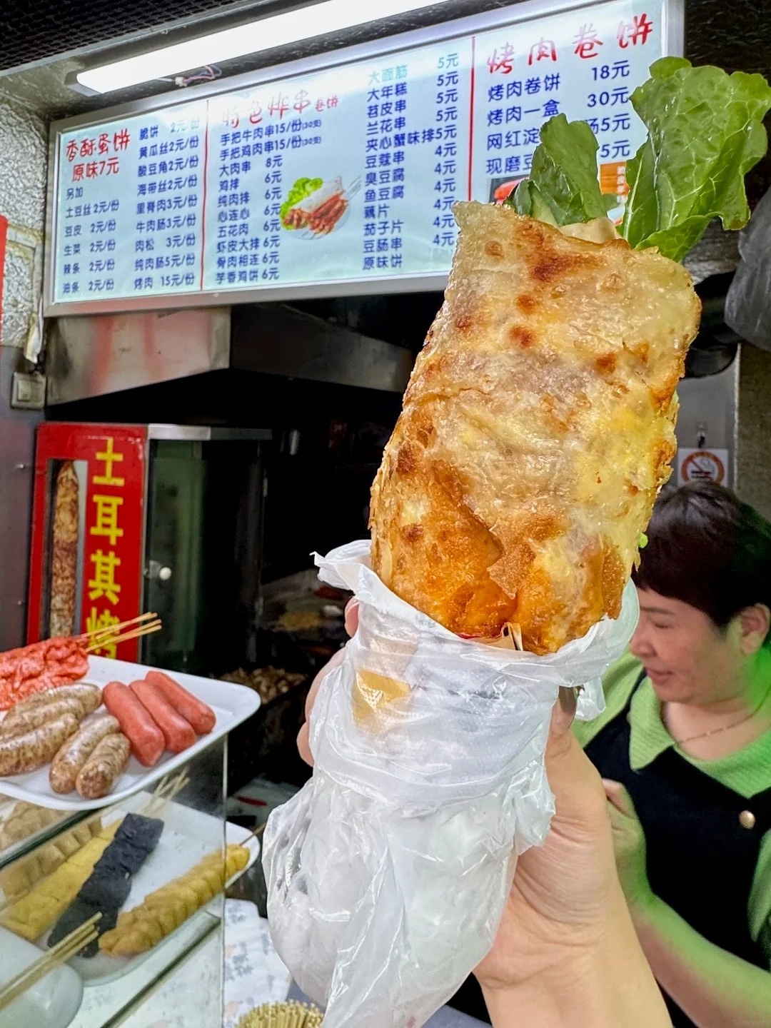 A hand holds a large, golden-brown scallion pancake wrap filled with lettuce and other ingredients, wrapped in a clear plastic bag. In the blurred background, a brightly lit menu board displays Chinese characters and prices, indicating a street food stall, with a woman in a green top visible behind it.