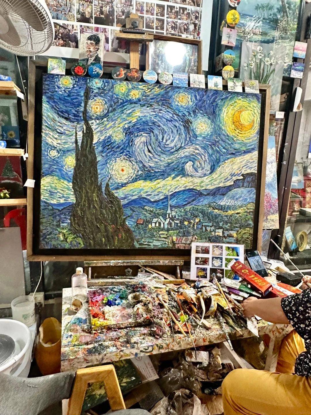 A large, textured oil painting of Van Gogh's Starry Night is displayed on an easel in an art studio. In front of the painting is a messy artist's palette covered in colorful paints and numerous paintbrushes. Various small artworks and art supplies are visible in the background and on shelves.