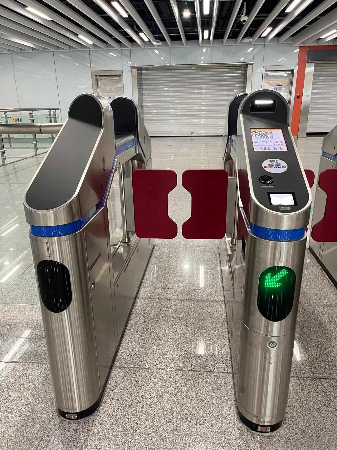 Two modern stainless steel metro turnstiles with red swing arms. The right turnstile has a touchscreen display showing various payment icons and instructions, including QR codes and a Visa logo. A green illuminated arrow pointing downwards is visible on the front of the right turnstile.