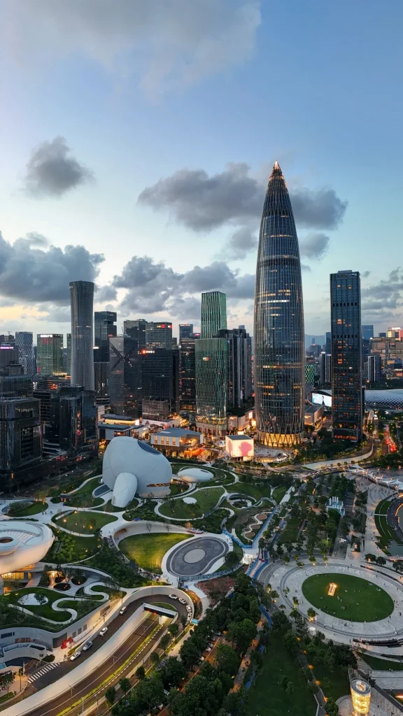 An aerial view of the Shenzhen skyline at dusk, featuring modern skyscrapers, including the prominent, needle-shaped Ping An Finance Centre. In the foreground, a contemporary urban park with curved pathways, green spaces, and distinctive white, organic-shaped buildings is visible. Roads with car traffic and illuminated city lights are also present.