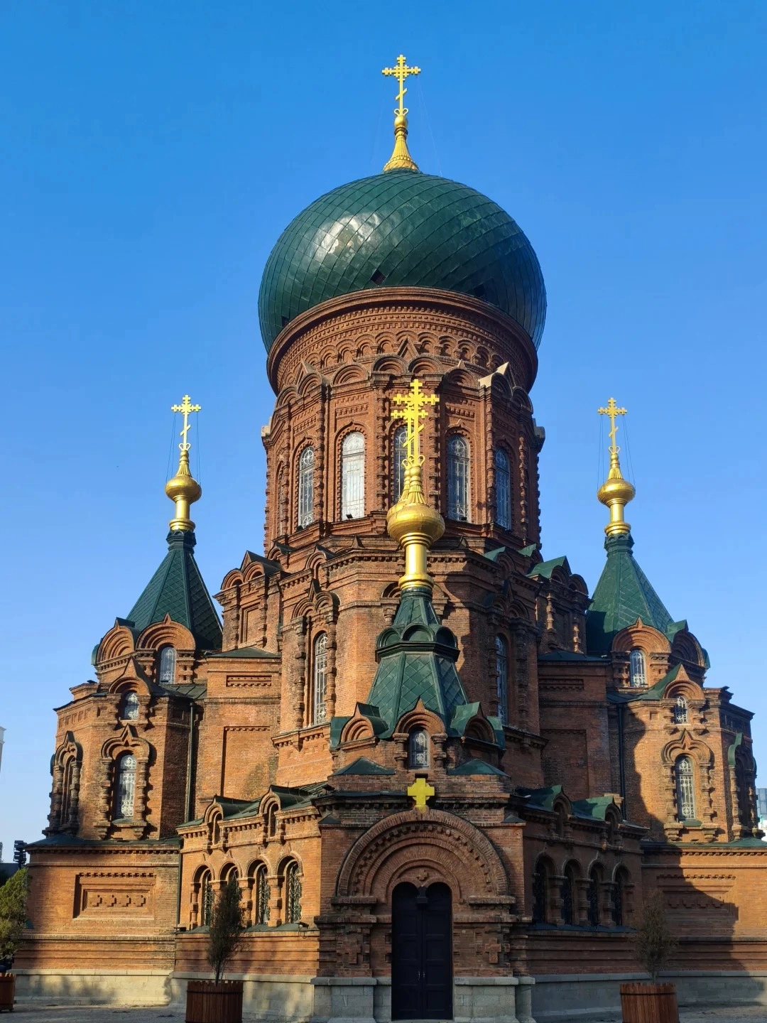 A large, ornate Orthodox cathedral constructed of red brick under a clear blue sky. It features a prominent, large green dome topped with a golden cross, and several smaller green conical roofs topped with golden crosses. The building has intricate brickwork, arched windows, and decorative elements.