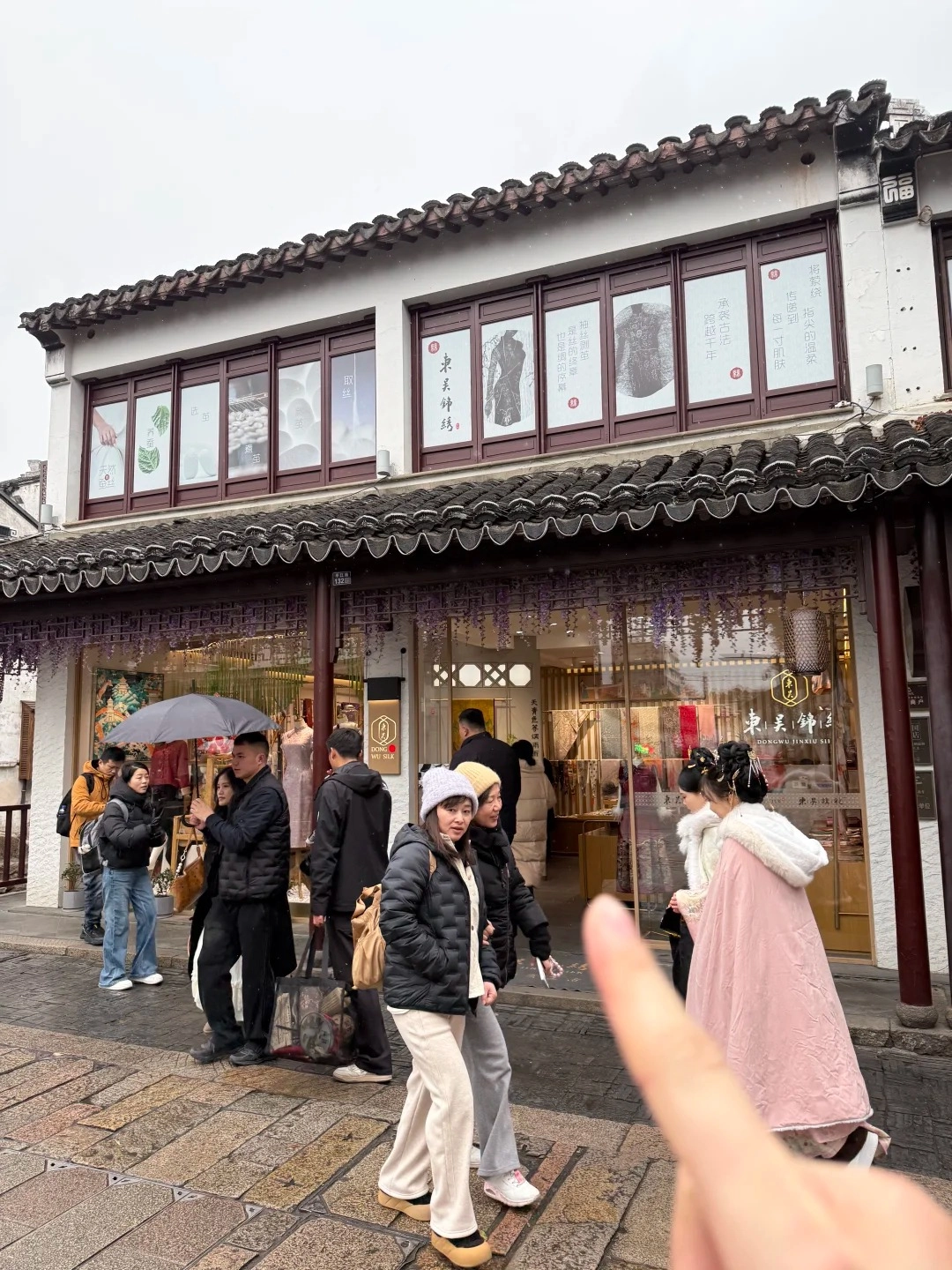 A group of people walk on a cobblestone street in front of a traditional Chinese building with ornate dark brown wooden window frames and tiled roofs. The building has multiple windows displaying artwork and fabric. Some people are carrying umbrellas, and it appears to be lightly raining or snowing. In the foreground, a person's hand is gesturing towards the building.