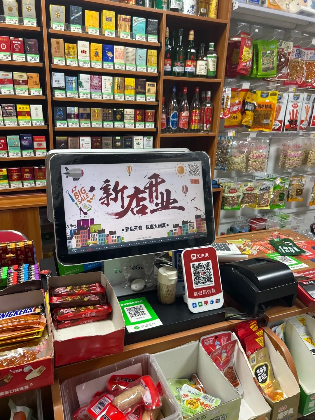 A digital screen on a counter in a convenience store displays 'New Store Opening' and 'Big Sale' in Chinese. To the right of the screen is a QR code for Alipay and WeChat Pay. Below the screen are boxes of snacks, candy bars, and cigarettes on shelves in the background.