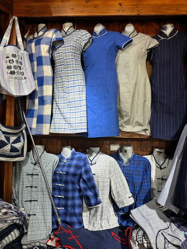 A display of traditional Chinese cheongsam dresses and jackets on mannequins and shelves. The garments feature various patterns like blue and white plaid, blue stripes, and subtle checkered fabrics. Some have mandarin collars and frog fastenings. A tote bag with a cat pattern hangs on the left, and a quilted fabric bag is also visible.