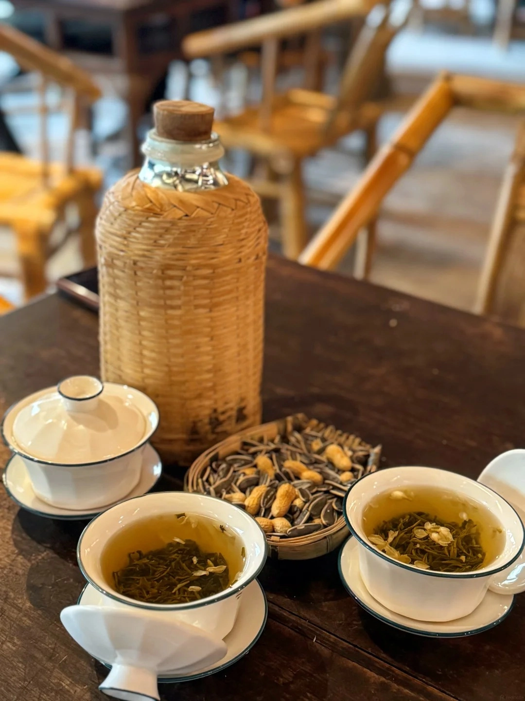A tall, woven bamboo bottle with a cork stopper sits on a dark wooden table next to two white gaiwan tea cups filled with green tea and a small woven basket filled with sunflower seeds and peanuts.