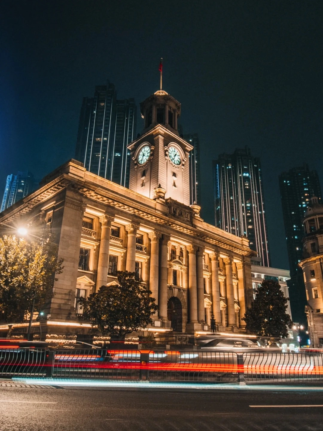 A grand, historic, European-style building with columns and a clock tower stands illuminated at night, with blurred red and white light trails from car traffic in the foreground and modern skyscrapers in the background.
