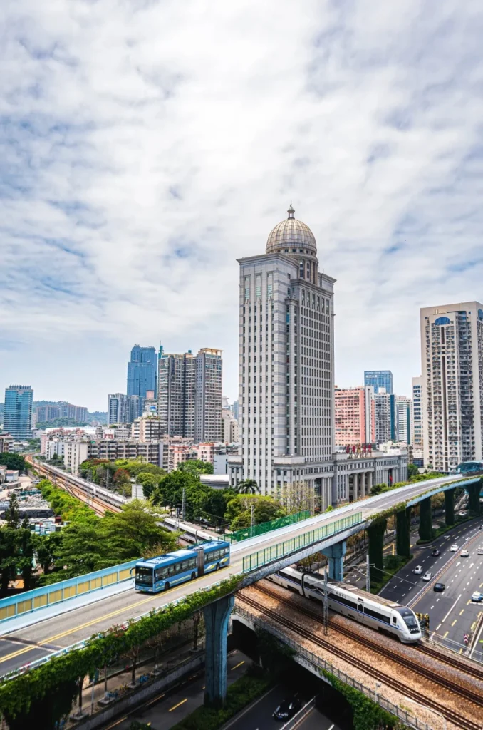 An elevated blue BRT bus travels on a dedicated lane above a multi-lane road and train tracks in Xiamen. A high-speed train is visible on the lower tracks. In the background, numerous modern skyscrapers and buildings form a dense cityscape under a cloudy sky. Green foliage covers the supports of the elevated busway.