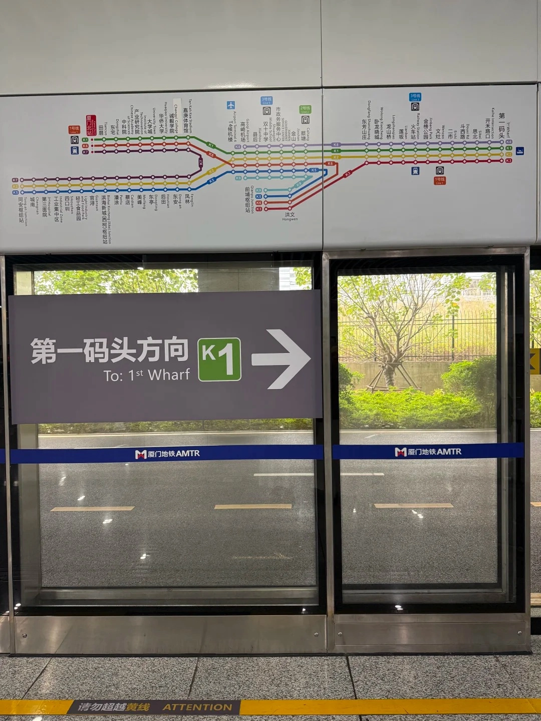A detailed metro system map is displayed on a white wall above a station platform. Below the map, a large sign with Chinese characters and the English text 'To: 1st Wharf' points to the right, indicating the direction of the K1 line. A glass door with a blue stripe displaying '厦门地铁 AMTR' is visible on the right side of the frame.