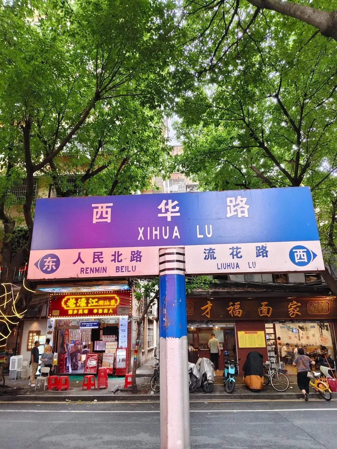 A street sign in Guangzhou, China, with text in Chinese characters and English. The sign reads 'Xihua Lu' in large English letters and below it 'Renmin Beilu' and 'Liuhua Lu'. The sign is purple and blue and is mounted on a striped pole. Below the sign, there are storefronts with illuminated signs and awnings, with people walking and eating. Trees with green leaves overhang the street.