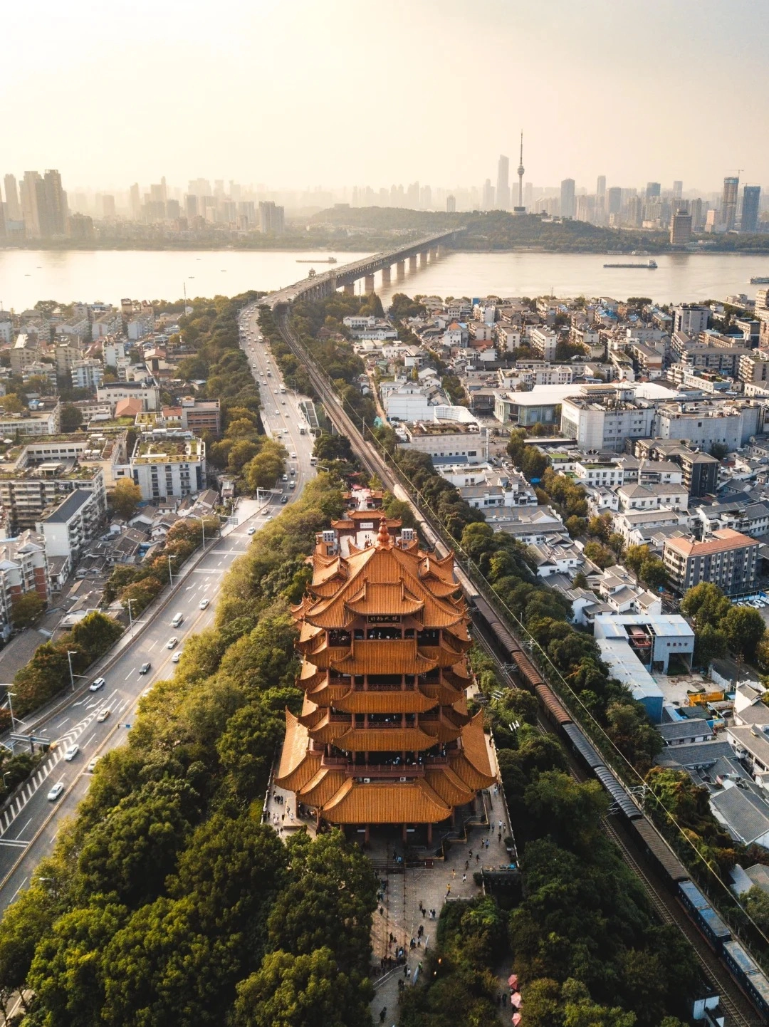 Aerial view of the Yellow Crane Tower, a multi-tiered traditional Chinese pagoda with a yellow-tiled roof, surrounded by lush green trees. A busy road with cars runs alongside the tower, and a railway track with a train is visible to the right. In the background, the Yangtze River flows, spanned by a long bridge, with the cityscape of Wuhan visible on the opposite bank, including a tall television tower.