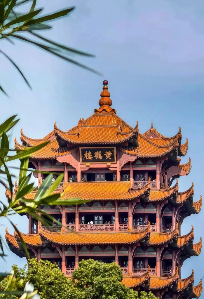The upper levels of the Yellow Crane Tower, a traditional Chinese multi-tiered pavilion with orange tiled roofs and red painted columns, are visible against a clear blue sky. Green foliage from trees and branches frame the lower and left sides of the image. A black plaque with Chinese characters is mounted on the central structure.