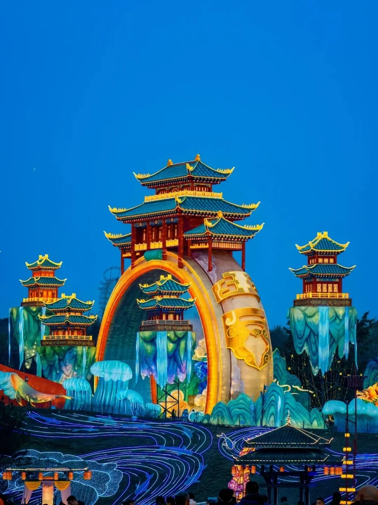 An elaborate nighttime lantern display featuring illuminated traditional Chinese-style temples with multi-tiered roofs and golden accents, set against a clear blue night sky. The lanterns are arranged on sculpted mountains and waterfalls made of light, with glowing blue rivers and pathways in the foreground.