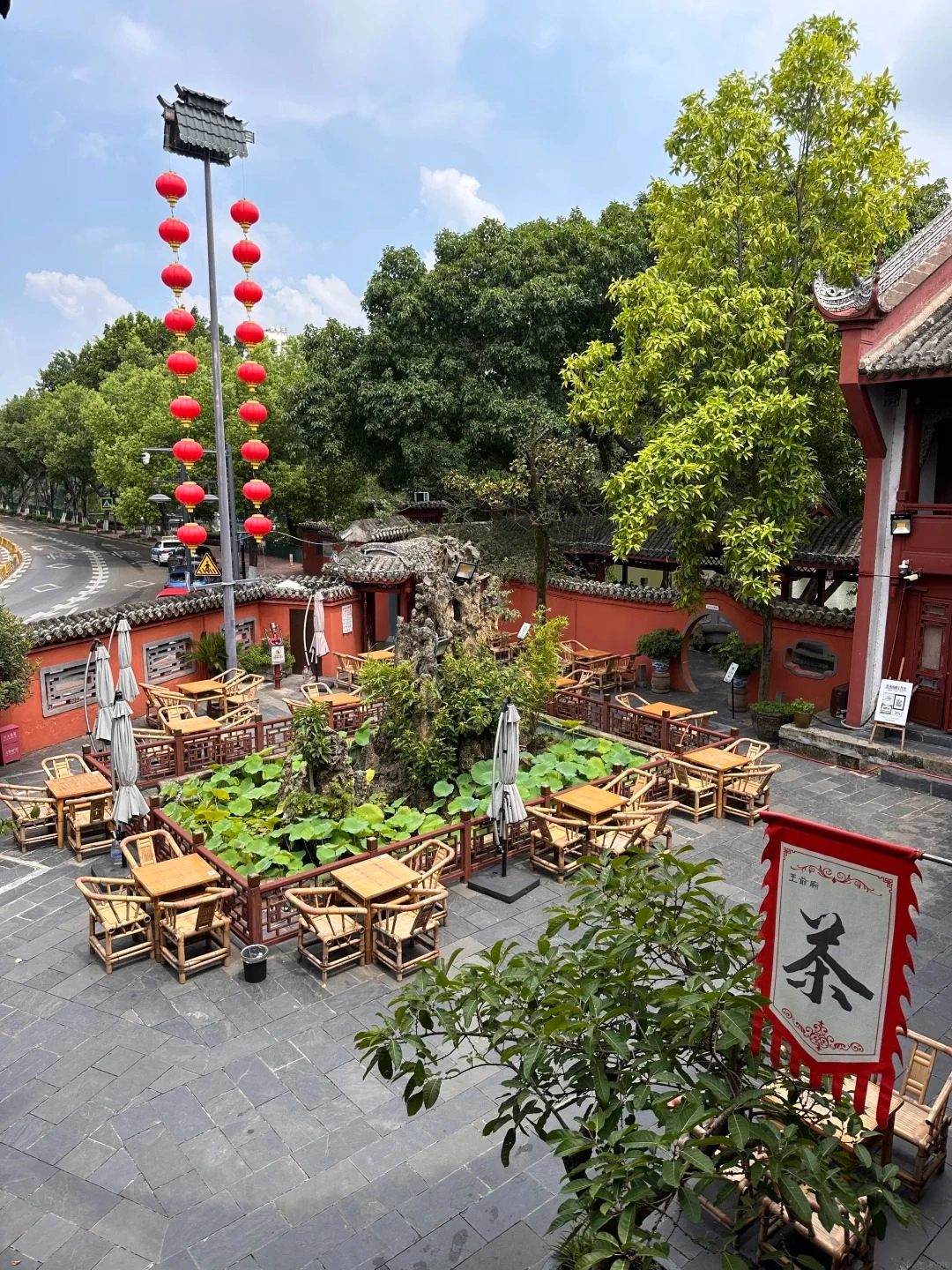 An outdoor courtyard with many bamboo tables and chairs set up for seating. In the center is a small pond with lotus plants and a rock feature. Red lanterns hang from a tall pole on the left side of the image. To the right, a red building with traditional Chinese architecture and a banner that says 'Tea' in Chinese characters.