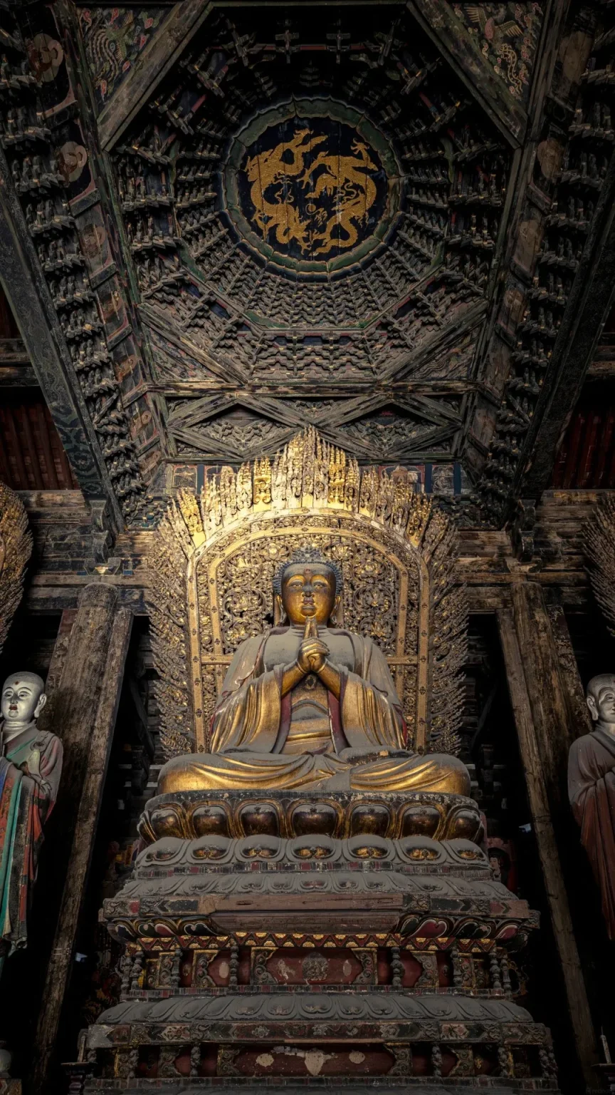 A large, golden Buddha statue sits on a tiered, ornate pedestal inside a dimly lit temple. The Buddha is in a meditative pose with hands clasped. Flanking the Buddha are two smaller, pale statues of monks. The ceiling above is intricately carved dark wood with a circular golden dragon motif in the center.
