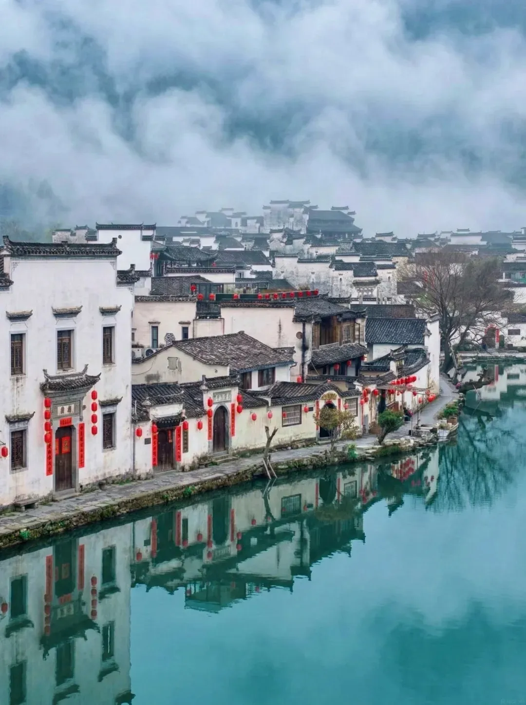 A serene village scene with traditional Chinese architecture featuring white-washed walls and dark tiled roofs, lining a tranquil waterway. Red lanterns are visible hanging outside some buildings. The buildings and surrounding mist are reflected in the still, turquoise water.