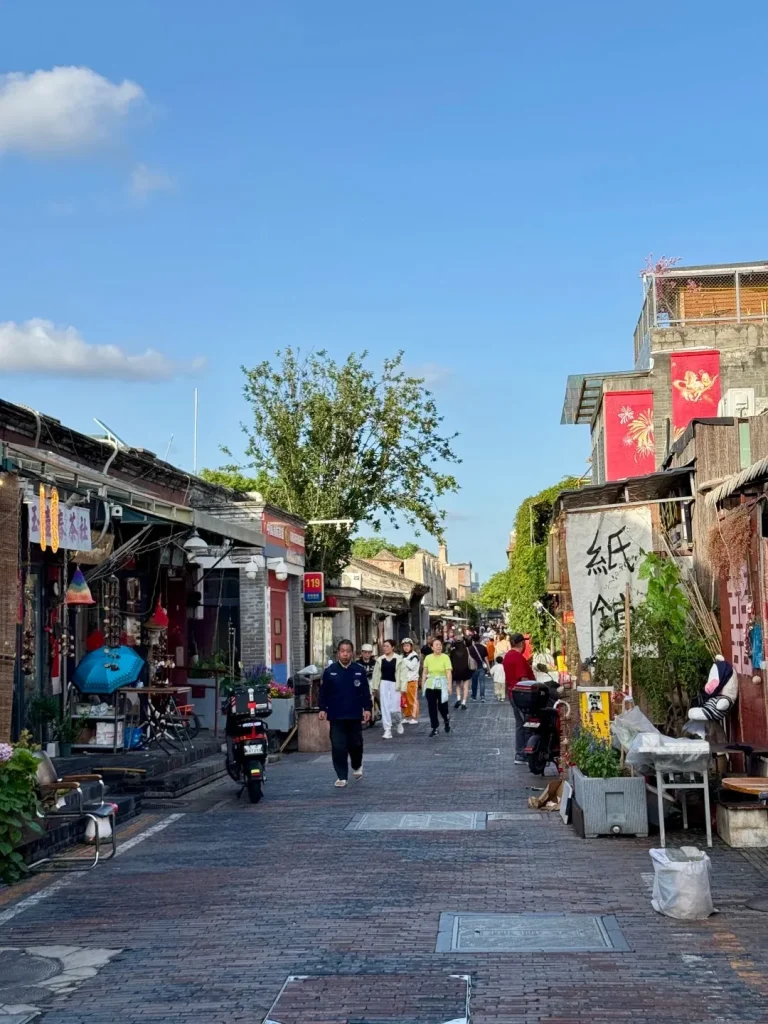 A view down a narrow, cobblestone alley lined with traditional two-story buildings on both sides. The buildings have grey brick facades and ornate rooftops, with many displaying colorful banners and signage. Shops are visible with merchandise displayed outside, including lanterns, umbrellas, and plants. Several people are walking down the alley, some browsing the shops. A tree with green foliage grows in the middle of the alley. The sky above is blue with scattered white clouds.