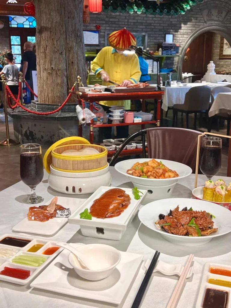 A chef in a yellow robe and a conical red and yellow hat is carving a roasted duck on a cart in a restaurant. Sliced Peking duck is served on white platters on a table with various dipping sauces and side dishes, along with glasses of dark liquid.
