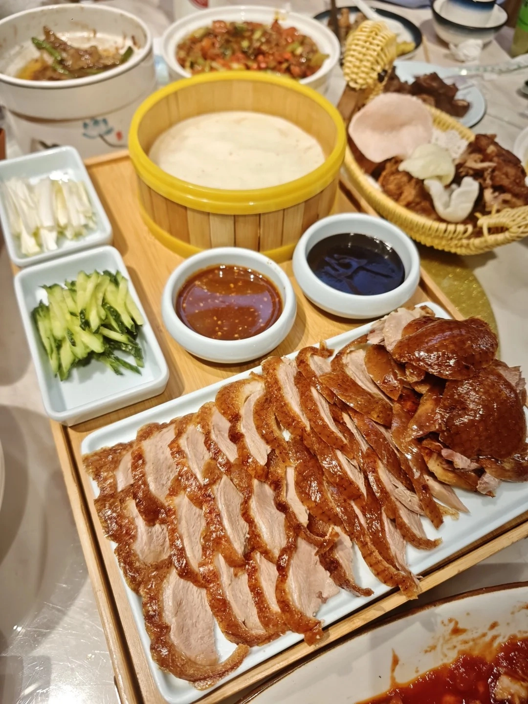 A long white platter filled with sliced Peking duck, featuring crispy brown skin and moist meat. To the left are small white dishes containing shredded cucumber and scallions, two dipping sauces (one dark, one reddish-brown), and a bamboo steamer basket filled with thin, pale pancakes. In the background are other dishes of food.