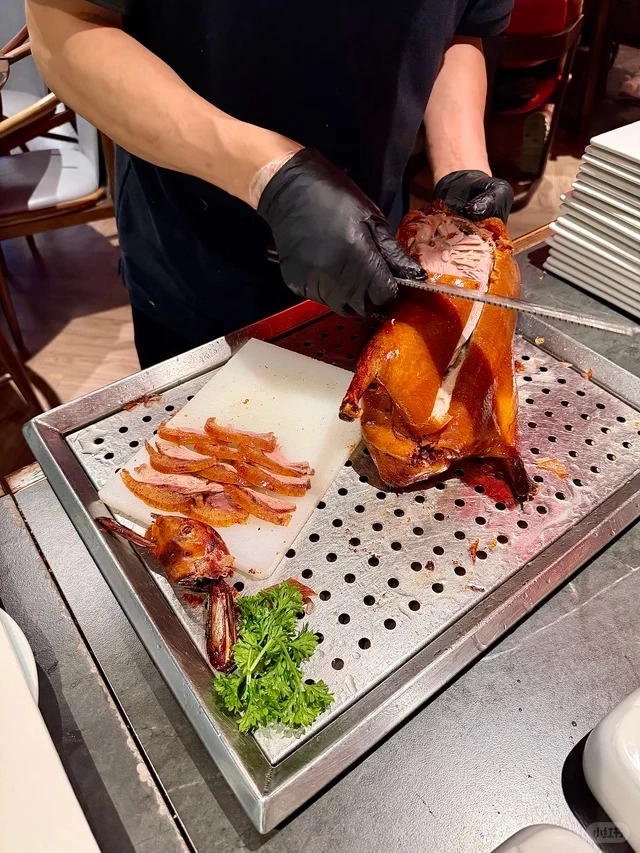 A chef wearing black gloves is carving a whole roasted Peking duck with a serrated knife. The duck is golden brown and glistening. Slices of duck skin and meat are already on a white cutting board. The carving is taking place over a metal tray with a perforated surface. A sprig of parsley is visible on the lower left. In the background, there are chairs and stacked plates.