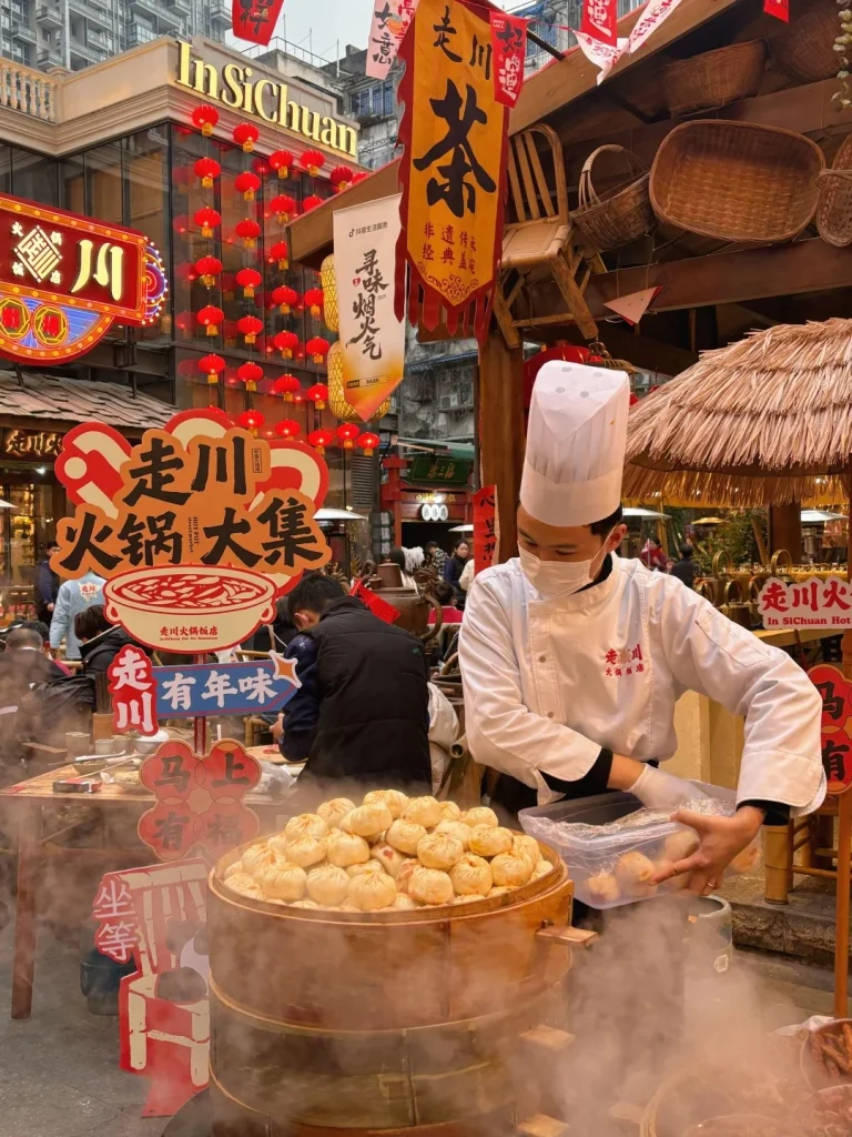 A chef wearing a white chef's hat and mask is serving steamed buns from a large bamboo steamer at an outdoor food stall decorated with red lanterns and banners. The steamer is filled with golden-brown, round buns, and steam is rising from it. In the background, there are signs with Chinese characters and other food stalls.