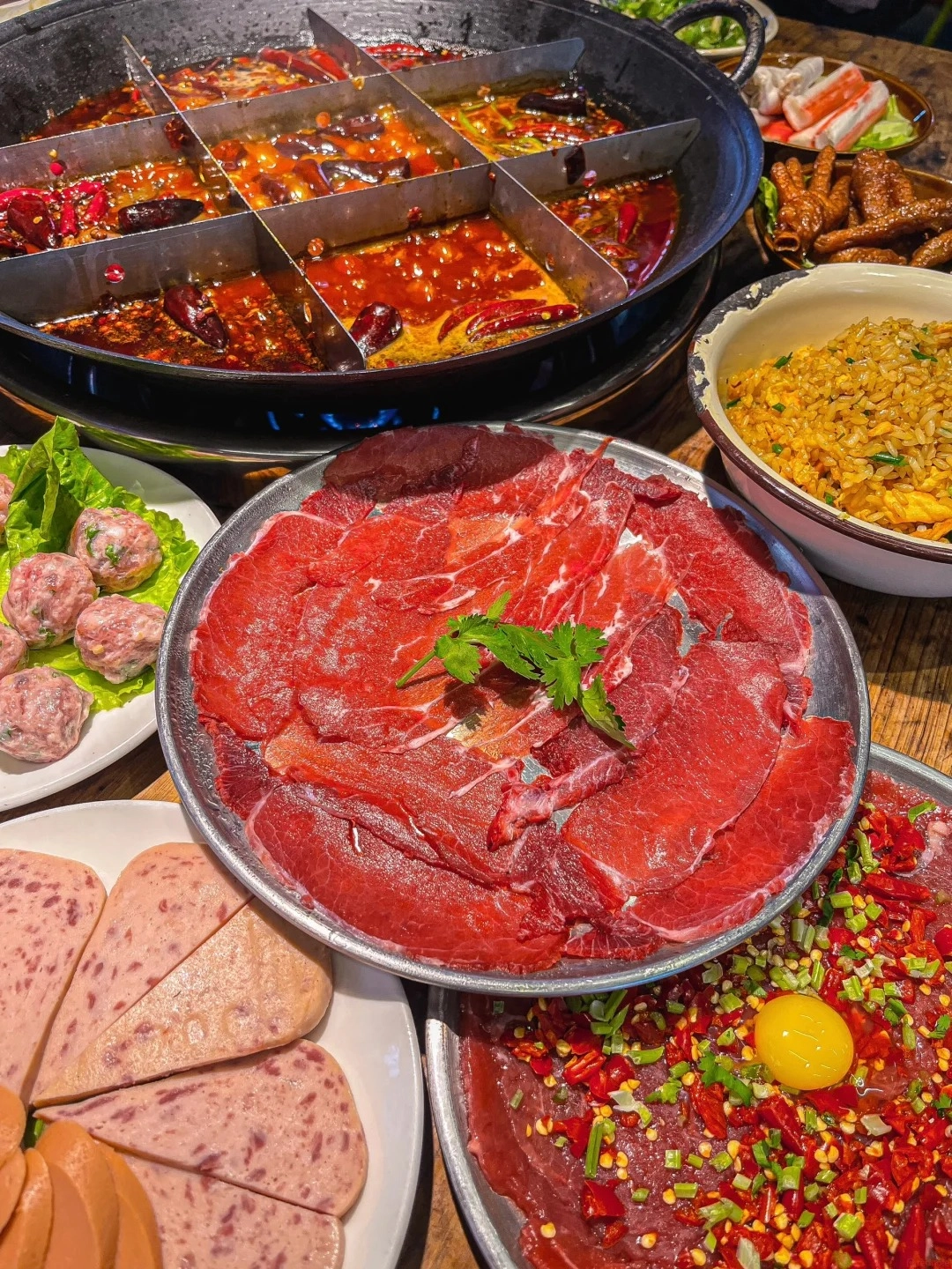A split-pot Sichuan hotpot with various broths and ingredients, a platter of thinly sliced raw beef, a plate of assorted sliced processed meats including ham and sausage, a bowl of fried rice, a plate of meatballs on lettuce, and a platter of chopped raw beef mixed with red chilies and a raw egg yolk.