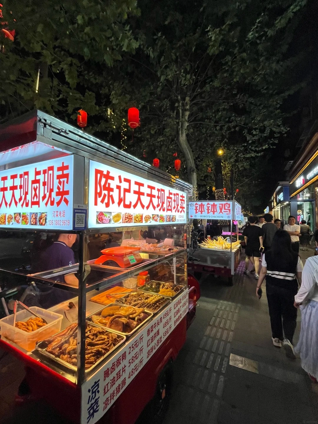 A brightly lit street food stall at night in Chengdu, displaying various prepared foods in metal trays. Chinese characters on signs advertise the food items. Red lanterns hang overhead, and a pedestrian walkway with patterned paving stones is visible.