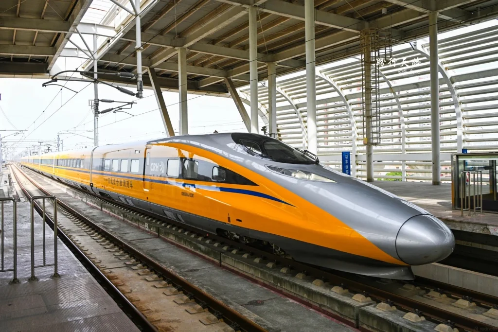 A silver and yellow high-speed train (CRH) with blue accents is stopped at a station platform under a modern, covered structure with visible support beams and overhead electrical lines. The train has multiple windows and a sleek, aerodynamic front. The tracks are visible in the foreground.