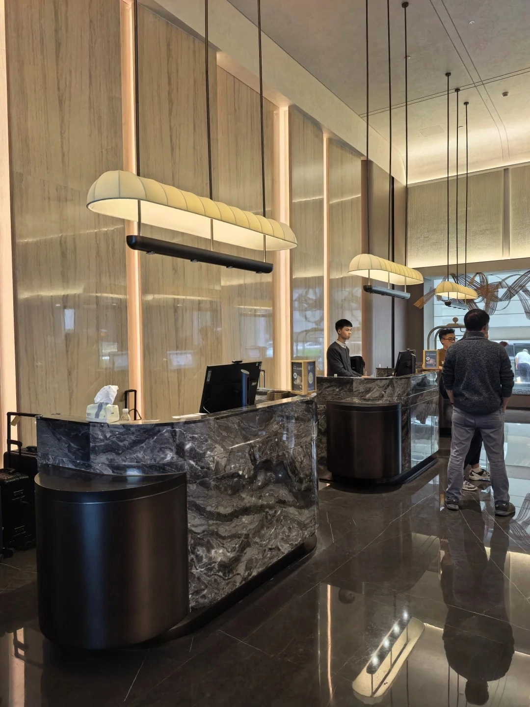 A modern hotel lobby in China with a marble front desk. A hotel employee stands behind the desk, and two guests are standing in front of it. A piece of luggage is visible on the left side of the desk.