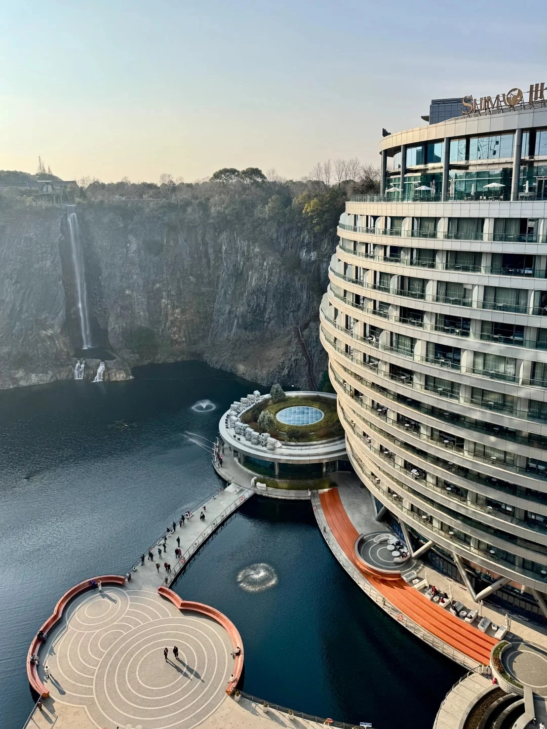 A modern, multi-story hotel with a curved facade built into the side of a quarry, overlooking a dark lake with a waterfall cascading down the quarry wall. A walkway connects the hotel to a circular building with a green roof and a glass dome, and a distinctively shaped outdoor seating area with concentric circles is visible on the left.