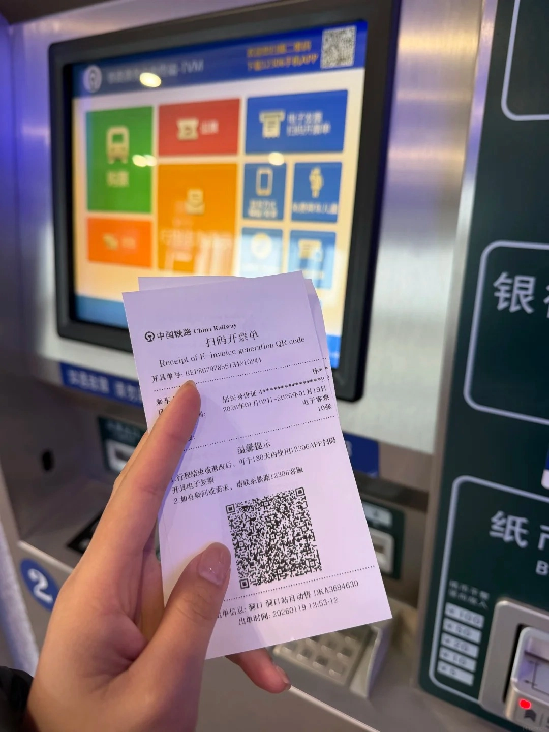 A hand holding a Chinese Railway e-invoice generation QR code receipt. In the background is a touch screen interface of a ticket machine with colorful icons.