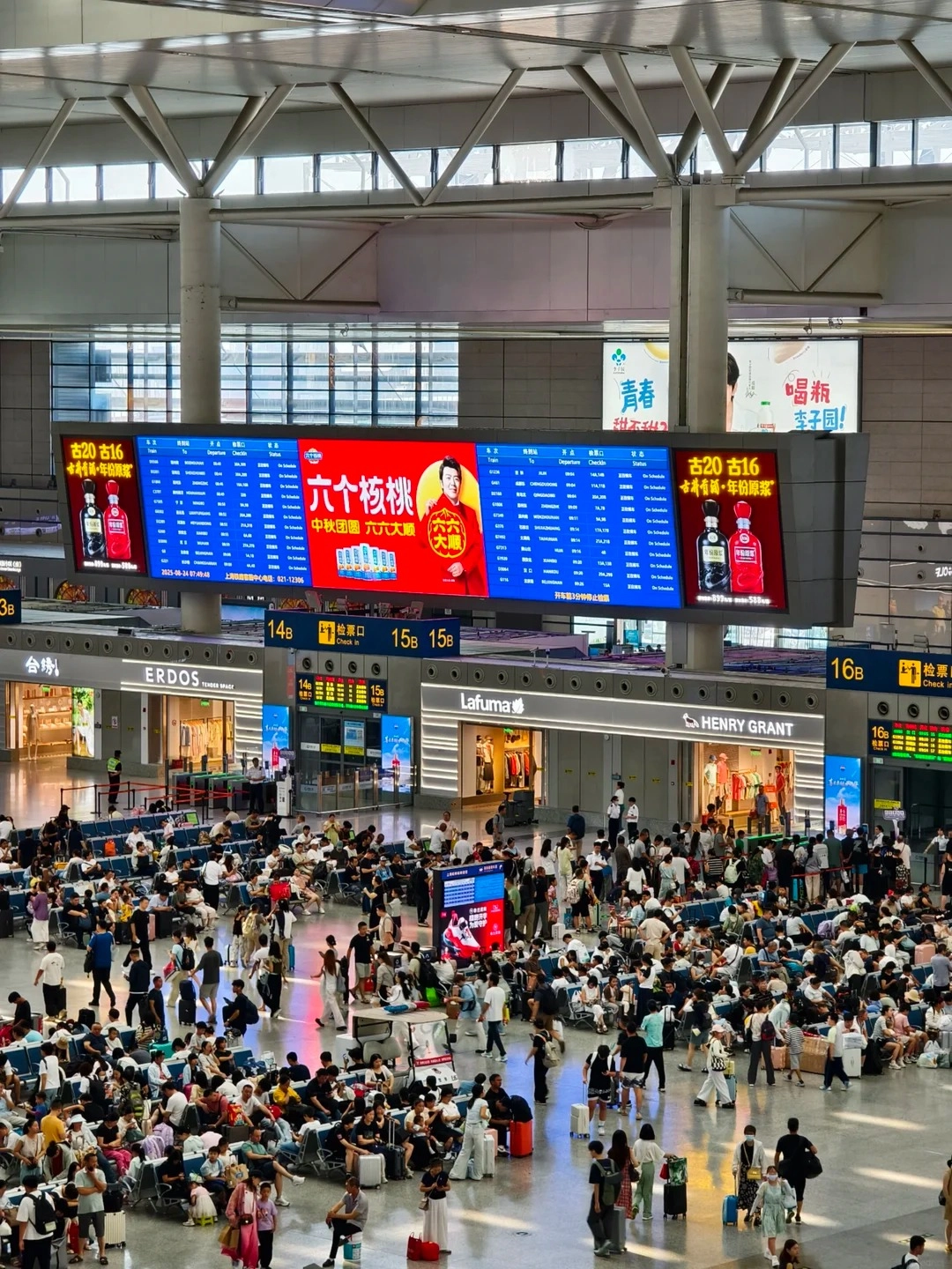 A wide shot of a bustling indoor train station concourse filled with many seated passengers and people walking. Above them, large, brightly lit digital departure boards display train information in Chinese and English, with advertisements for products and services. Overhead, the station's modern architecture with exposed steel beams and skylights is visible. The scene captures the scale and activity of a major Chinese transportation hub.
