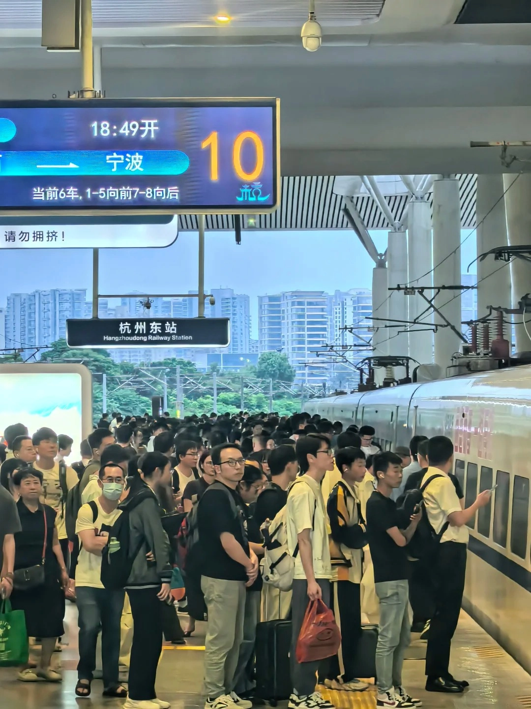 A crowded platform at a Chinese railway station, with a digital display showing train departure information and destination 'Ningbo' and platform '10'. Many passengers are standing with luggage, waiting to board a train.