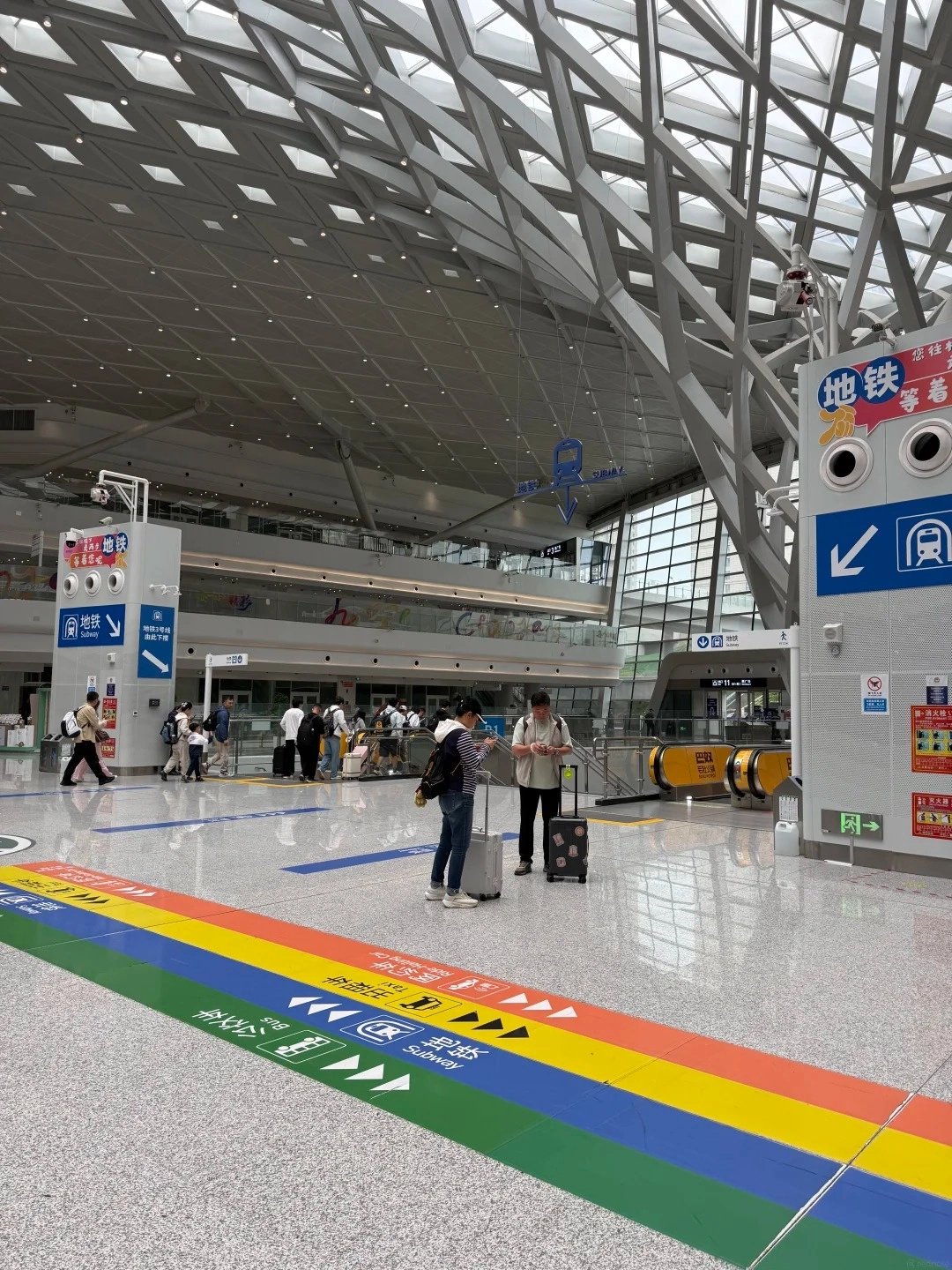 A wide-angle view of a modern, spacious train station interior with a high, arched ceiling and a prominent, gray, lattice-like structure. In the foreground, a colorful rainbow-striped floor guide indicates directions for different modes of transport, including subway, bus, and taxi. Several travelers with luggage are visible walking and standing in the station. Signs with Chinese characters and pictograms are posted on pillars and walls, directing passengers.