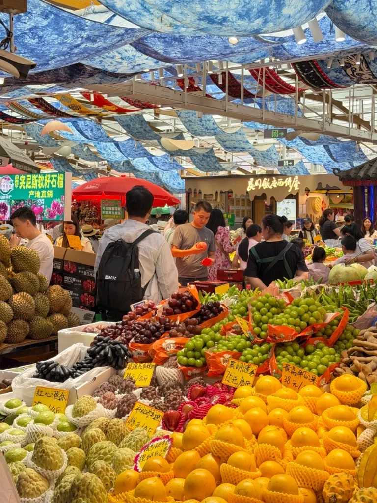 A bustling outdoor market in China with a variety of fresh fruits on display. In the foreground, there are large piles of durian, grapes (red and green), custard apples, mangoes, and other tropical fruits. People are browsing the stalls. Colorful fabric decorations hang from the ceiling, creating a canopy over the market. Some signs in Chinese characters are visible near the fruits.