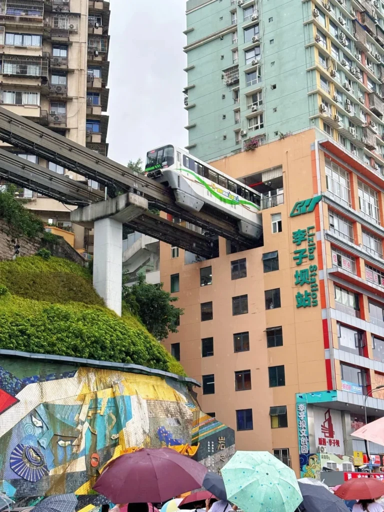A monorail train passing through a residential high-rise building above a street with colorful umbrellas and a mural on a retaining wall.