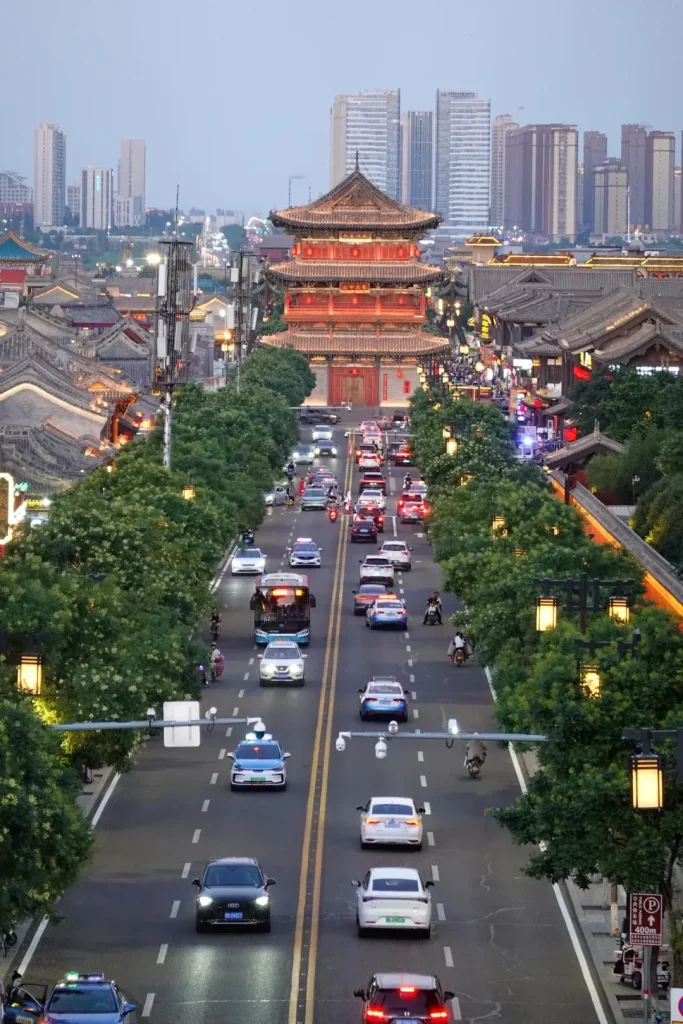 A wide, tree-lined road with heavy traffic leading towards a large, ornate, multi-tiered traditional Chinese temple building. The temple is brightly lit with red and gold accents. On either side of the road are traditional Chinese buildings with tiled roofs, some with lit lanterns. In the background, modern high-rise buildings form the city skyline under a pale twilight sky. Streetlights and traffic lights are visible, illuminating the vehicles and the road.