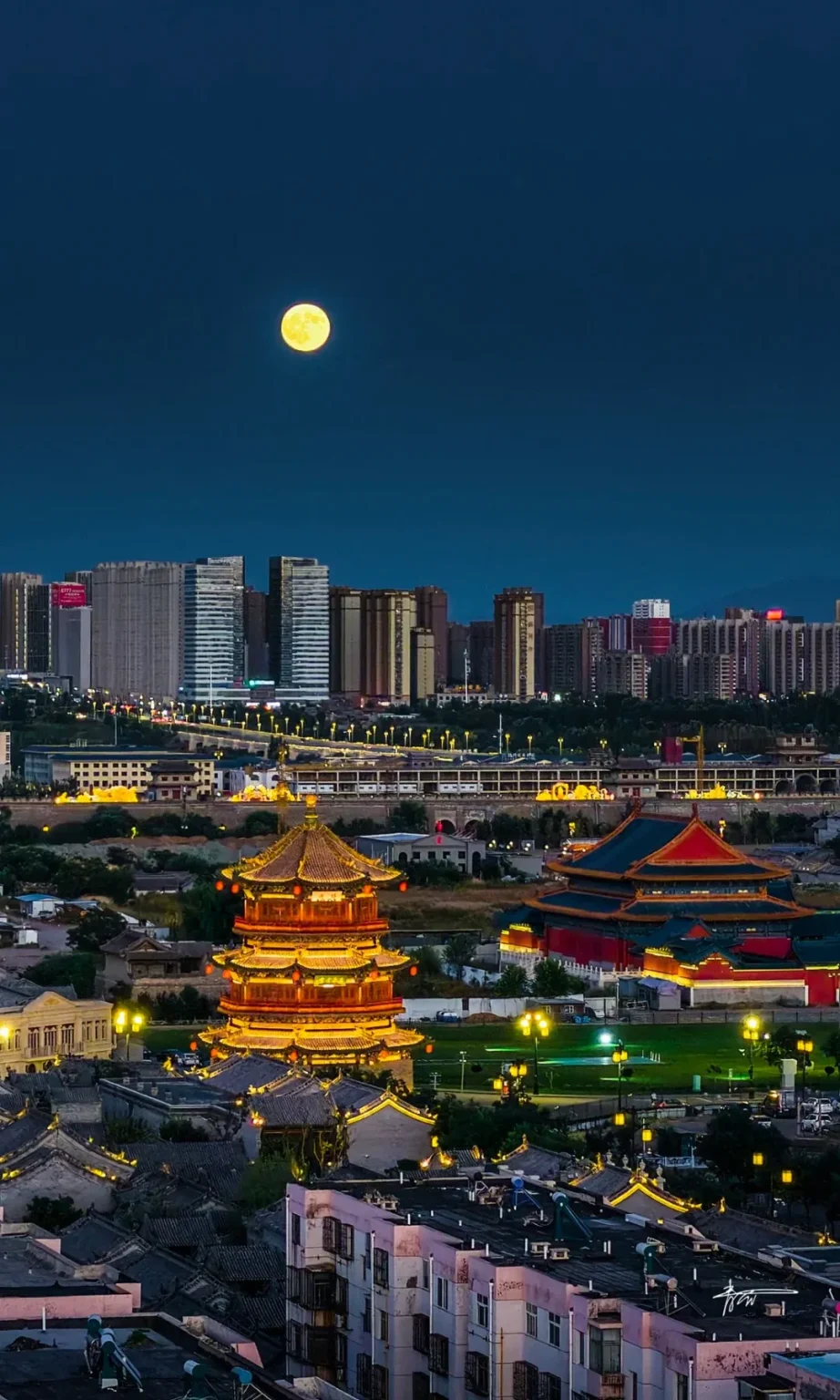 A panoramic night view of Datong, China, featuring a brightly lit ancient multi-tiered pagoda in the foreground. To the right of the pagoda is a traditional red and blue roofed building complex. In the background, a cluster of modern high-rise buildings stands under a dark blue night sky with a prominent, glowing full moon.