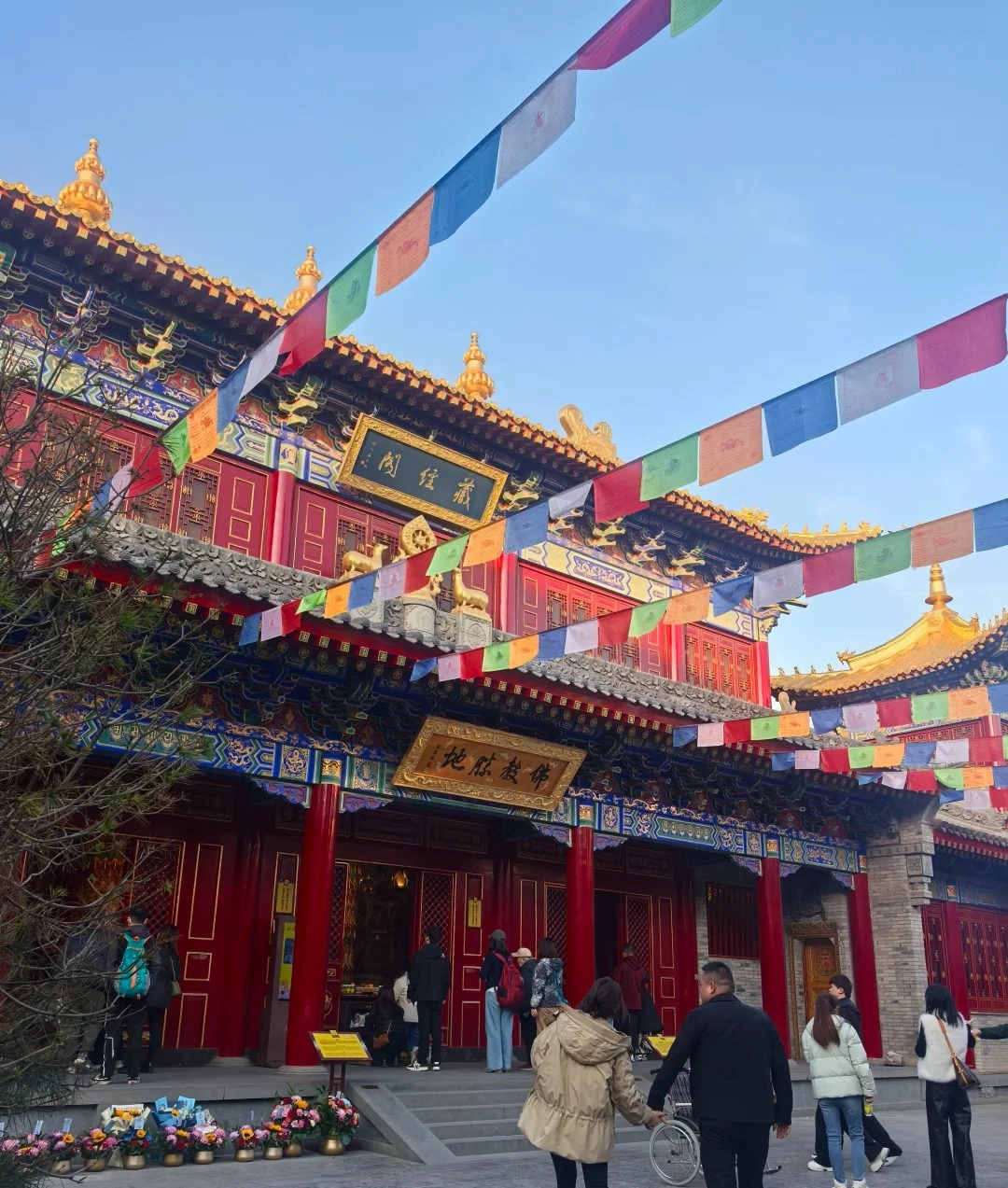 A wide-angle view of the exterior of the Guangren Tibetan Buddhist Temple in Xi'an, with colorful prayer flags strung across the front and bright red doors and golden decorative elements on the building's facade.