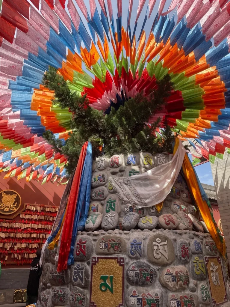 A close-up view of a stupa decorated with prayer stones, featuring Tibetan script. Colorful prayer flags fan out from the top of the stupa against a blue sky. Colorful prayer flags are strung across the top of the image, creating a vibrant canopy. The stupa is constructed with stones, many of which are inscribed with Tibetan Buddhist symbols and mantras. Prayer scarves in red, blue, yellow, and white are draped over the stupa. A small, golden circular emblem with a wheel of dharma symbol is visible on a red wall to the left, alongside rows of small red prayer tags.