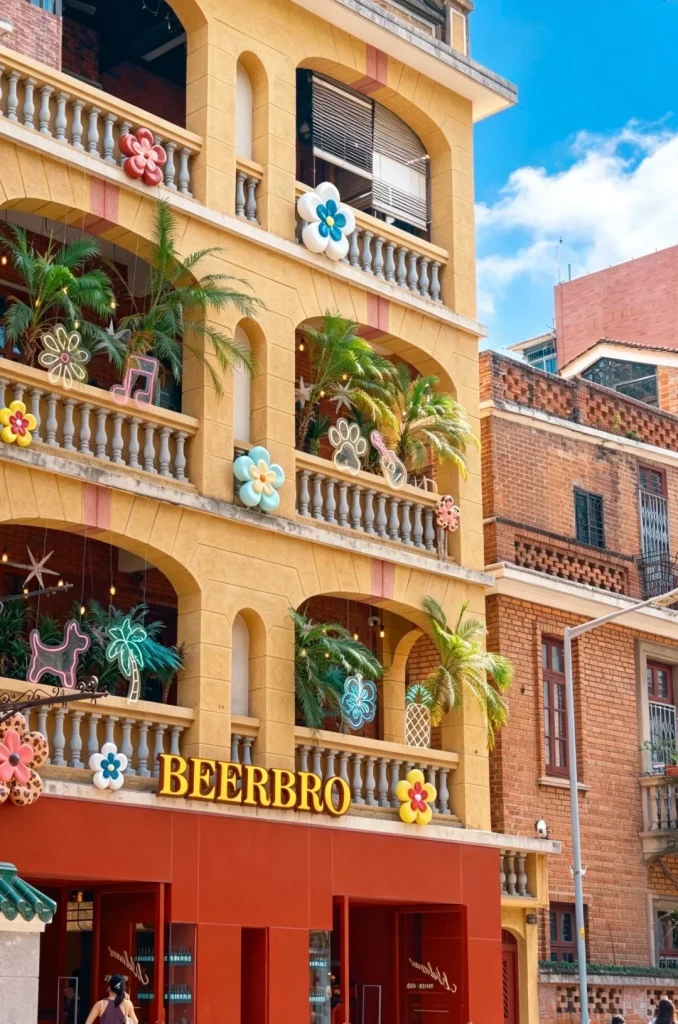 A multi-story building with a yellow facade featuring arched balconies decorated with colorful flower and neon light ornaments. The building has a red awning with the yellow sign 'BEERBRO' in the center. Palm trees and other greenery are visible on the balconies. To the right, a red-brick building with windows and balconies is partially visible under a blue sky with clouds.