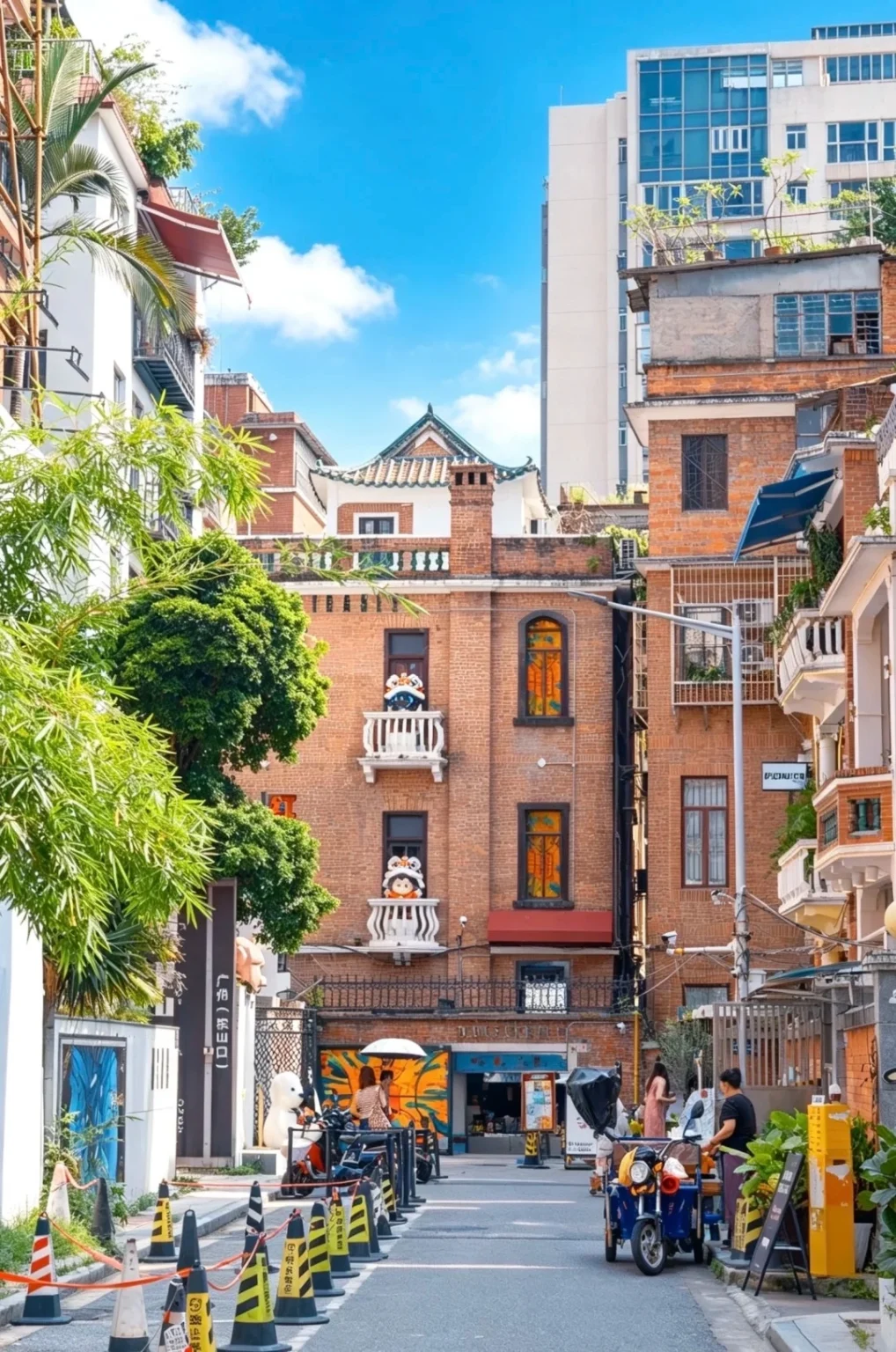 A narrow street in Guangzhou lined with red brick buildings and green trees under a blue sky with white clouds. Balconies on the buildings feature decorative figures. A blue scooter and people are visible on the street, with traffic cones and signage.