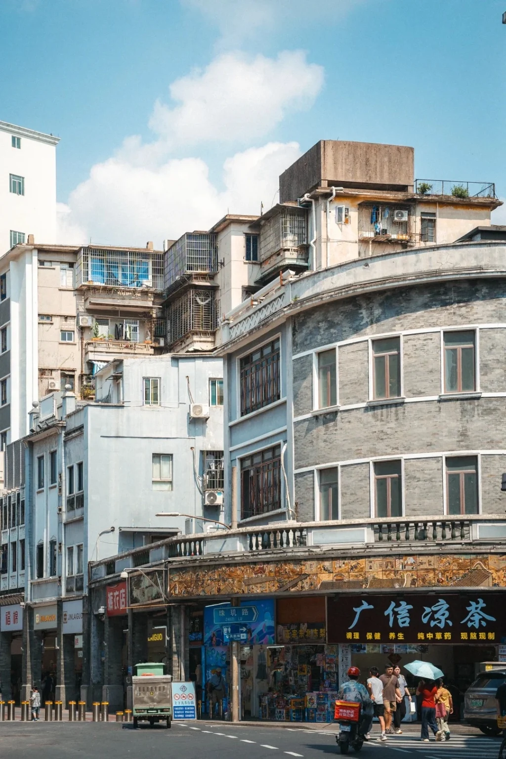 A multi-story street scene in Guangzhou, China, featuring traditional Qilou arcade buildings with their characteristic covered walkways and ornate facades. Several pedestrians are visible on the sidewalk, along with a street cart and a scooter. The buildings showcase a mix of architectural styles, with some featuring grey brickwork and balconies, while others are painted in lighter colors. The sky above is blue with scattered white clouds.