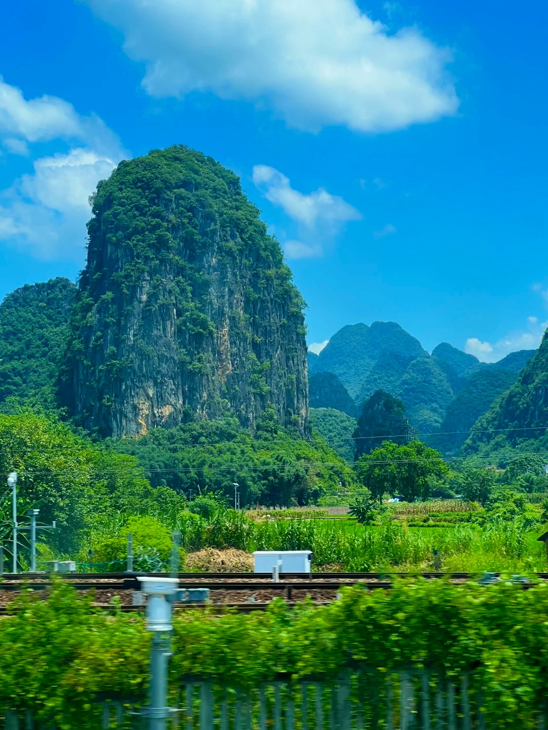 A large, steep, and verdant karst mountain dominates the center of the image under a bright blue sky with scattered white clouds. In the foreground, train tracks run horizontally across the frame, with lush green foliage in the immediate foreground and middle ground. Beyond the tracks, fields of green crops and scattered trees are visible, with more rolling green karst mountains in the background.